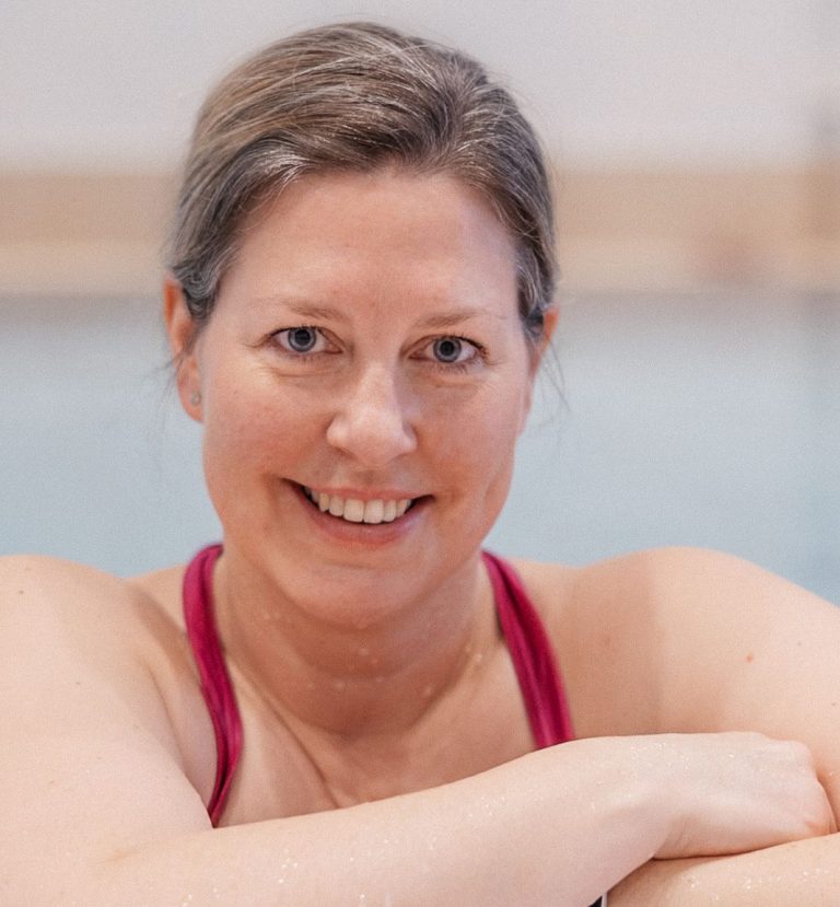 Portrait Foto Gründerin Eine lächelnde Frau, die im Wasser steht und einen pinken Badeanzug trägt.